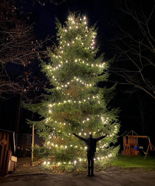 A professional holiday lighting installer posing in front of a stunning Christmas tree.